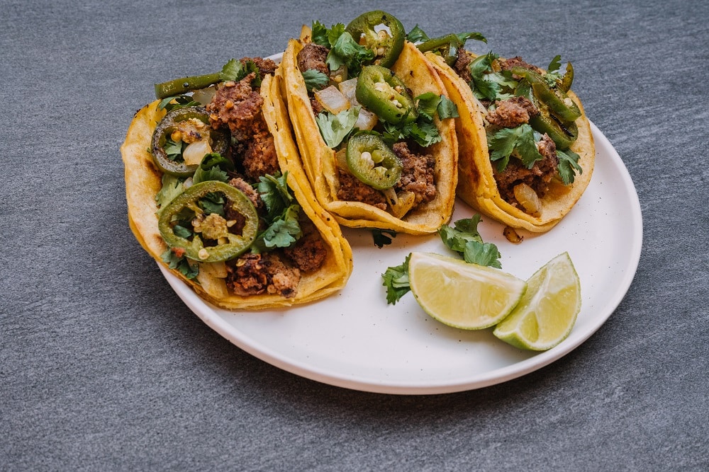 Three beef tacos topped with jalapeños and cilantro are served on a white plate with lime wedges, perfect for your next Taco Catering event.