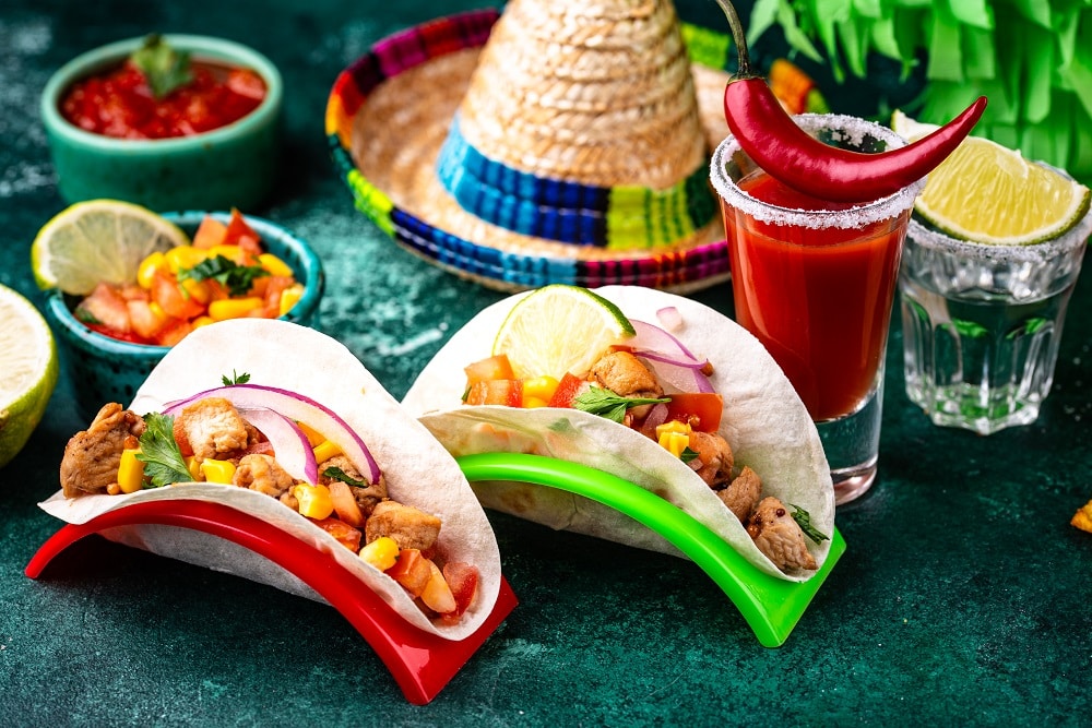 Two chicken tacos with vegetables in taco holders, a glass of cocktail with a chili pepper, lime wedges, salsa, and a sombrero hat in the background create an inviting Mexican vibe.