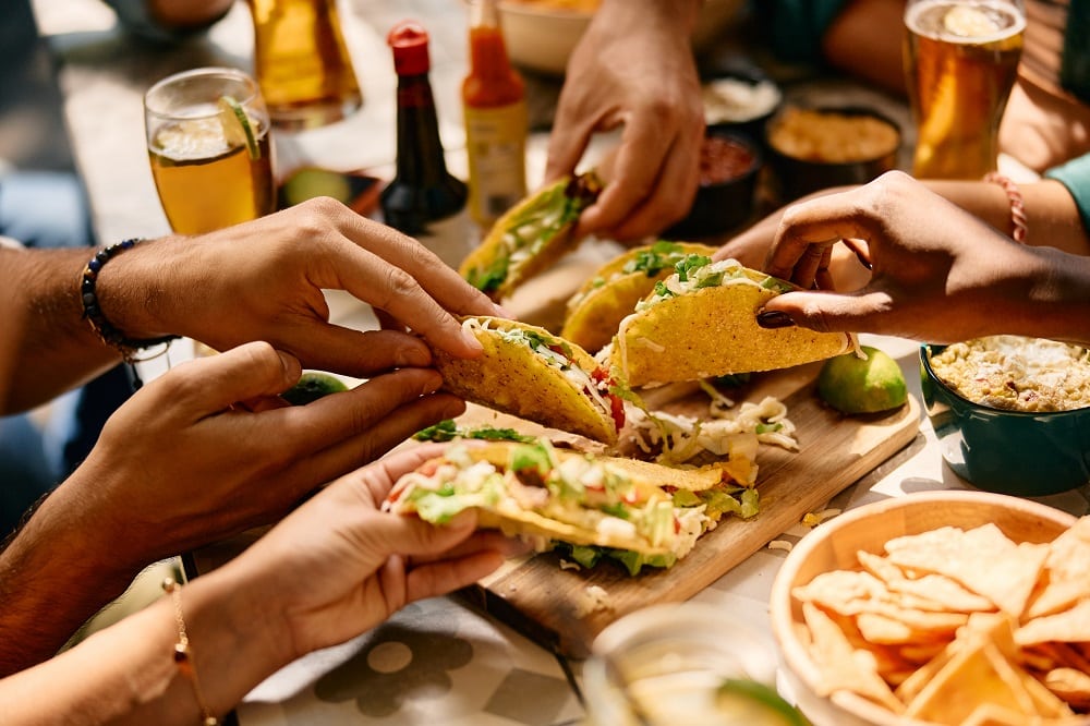 Several people reach for hard shell tacos at a table set with drinks, sauces, tortilla chips, and other delicious items from mexican catering.