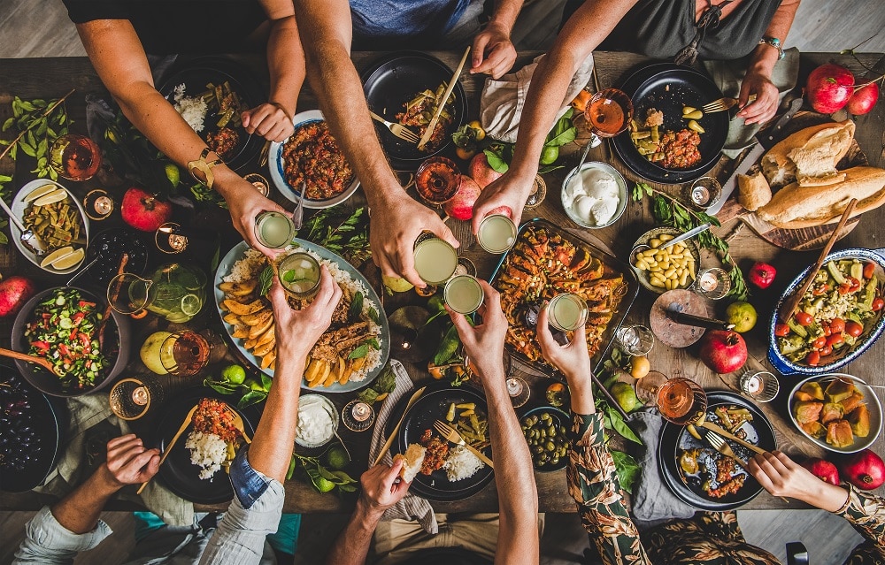 A group of people seen from above clinking glasses over a table filled with various dishes, salads, bread, and fruit.
