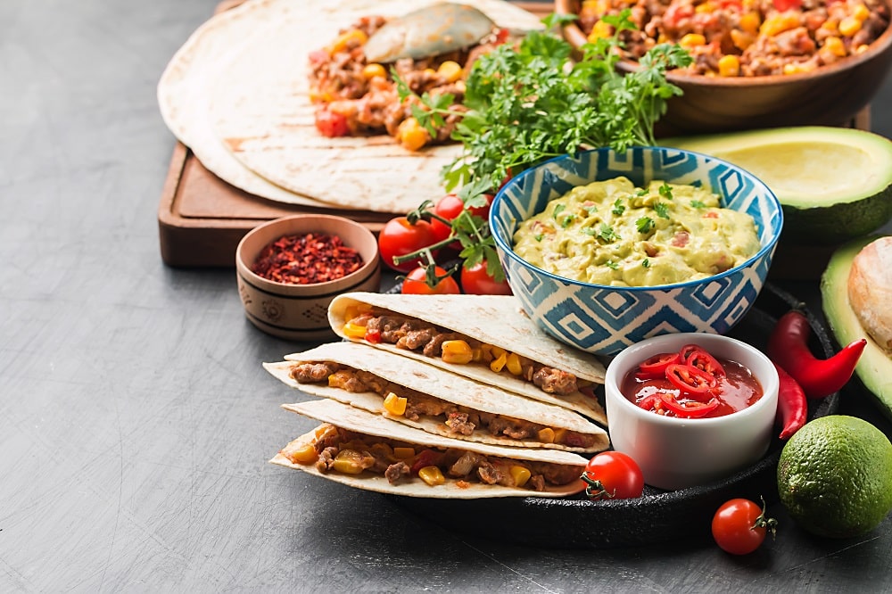 Three folded Mexican quesadillas with corn and beans, served with a bowl of guacamole, salsa, cherry tomatoes, avocado, and herbs on a gray surface.