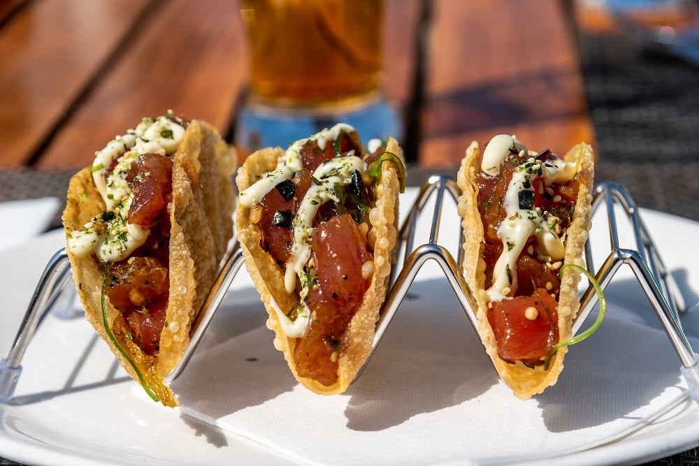 Three corn tortillas filled with shredded meat, sliced vegetables, and lime wedges sit on a white plate, perfect for comparing a DIY Taco Bar vs. Hiring a Taco Caterer for your next event. Served alongside are toasted bread slices and fresh salad.
