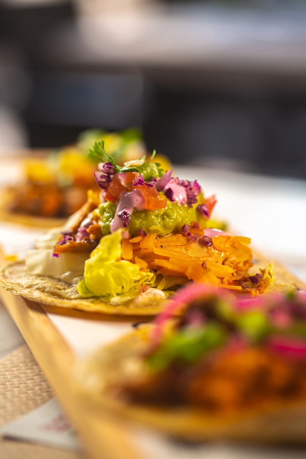 A close-up of open-faced tacos topped with shredded vegetables, guacamole, and garnishes on a wooden tray—perfect inspiration for your next gathering when choosing between a DIY Taco Bar vs. Hiring a Taco Caterer.