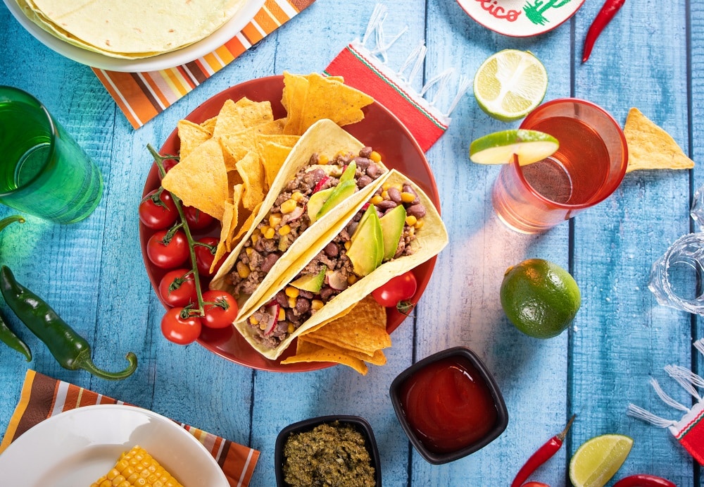 A plate with two tacos filled with beans, corn, and avocado, surrounded by tortilla chips and cherry tomatoes, sits on a blue wooden table with drinks, lime, and salsa—perfect for any Taco Catering event.